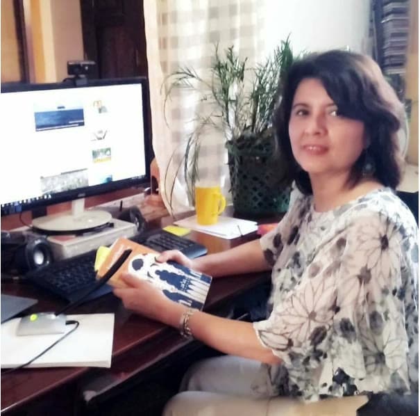 Amalia at her desk giving a lesson by video call, holding a Spanish textbook, with a plant and a yellow mug beside the monitor