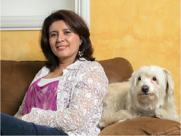 Amalia Jarquín smiling on a brown sofa next to her white dog, in front of a yellow wall
