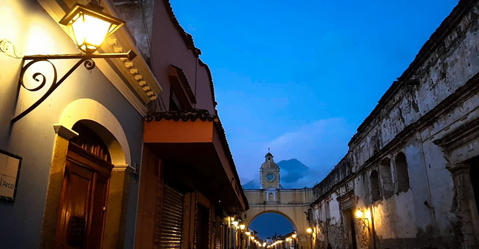 Calle del Arco in Antigua Guatemala at dusk: the yellow Santa Catalina arch, street lamps lit, the Agua volcano behind