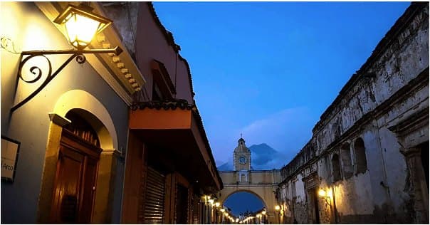 A street in Antigua Guatemala at dusk, with the Santa Catalina arch and the Agua volcano in the distance